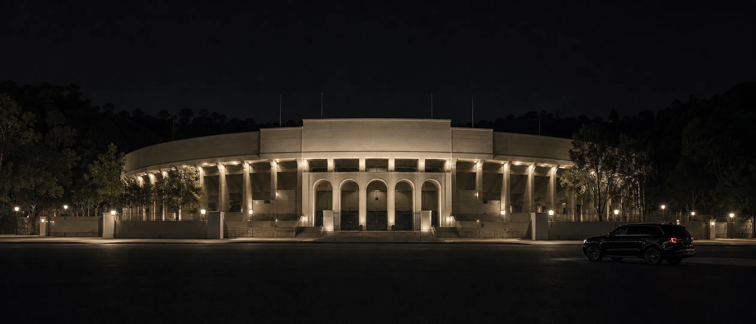 A near-black luxury SUV waits on the quiet approach to Pasadena's illuminated Rose Bowl at night.