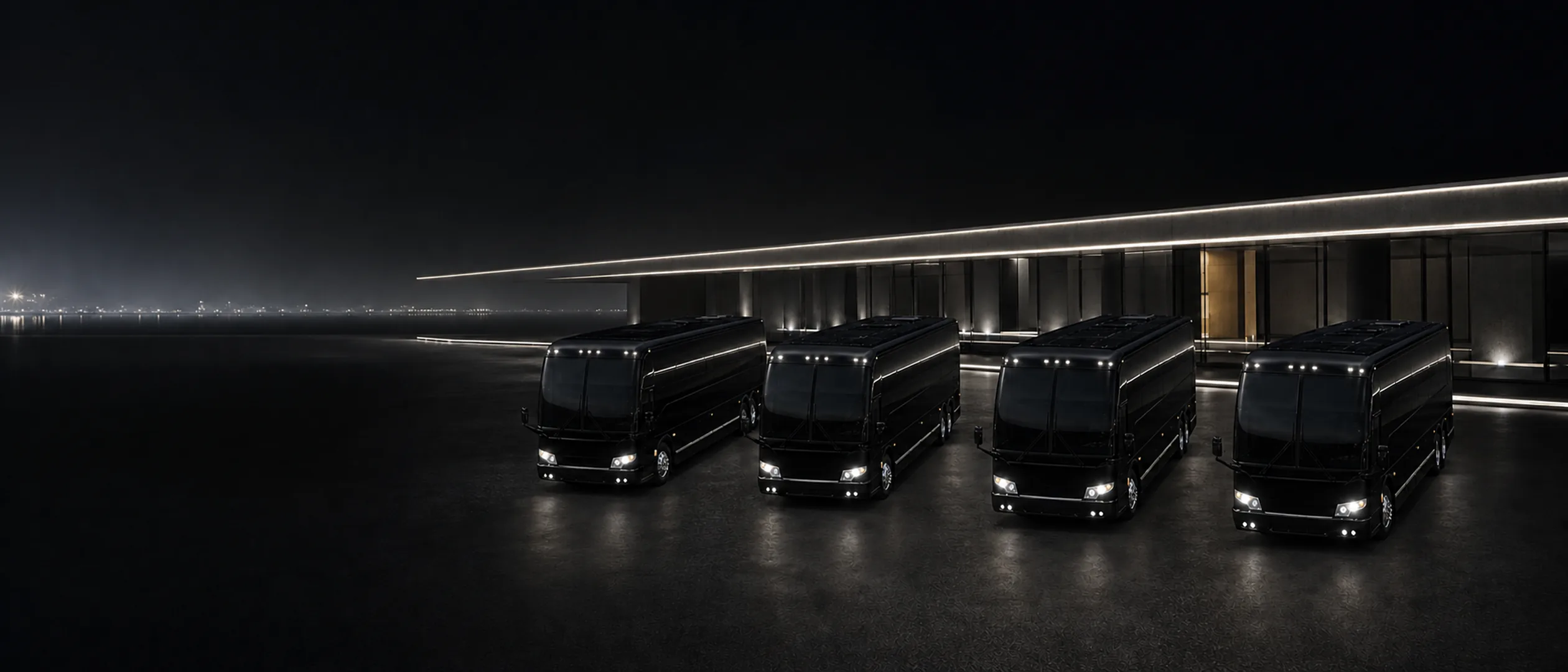 Four black Prevost H3-45 motorcoaches parked nose-out in a fanned arc at night, with a foggy waterfront and city lights beyond.