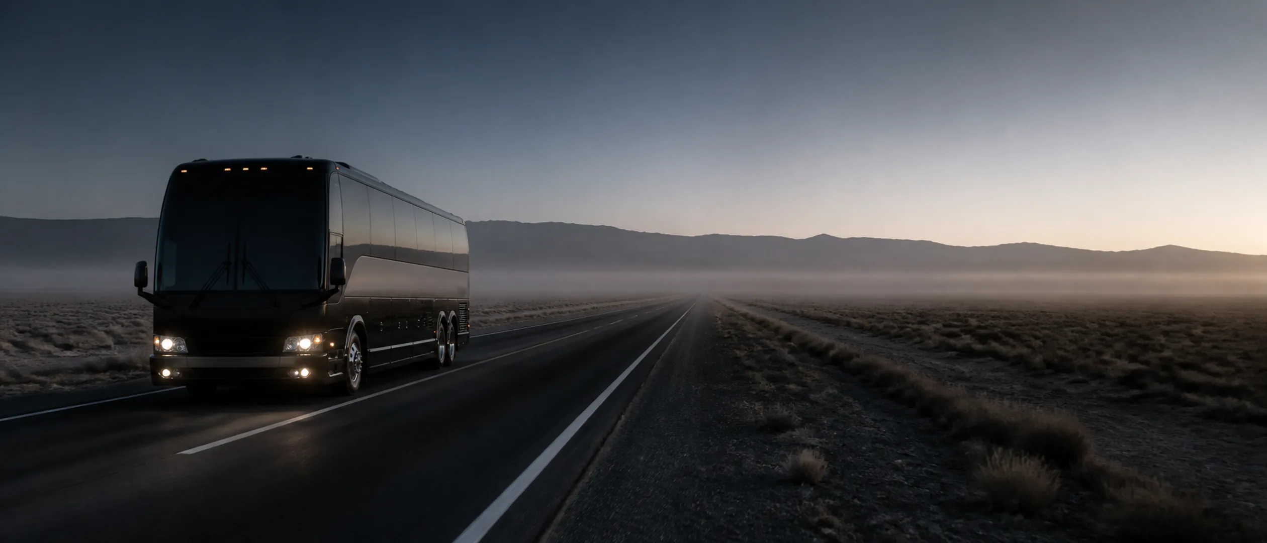 A black motorcoach with a tag axle drives down a two-lane desert highway at dusk, mountains in the distance.